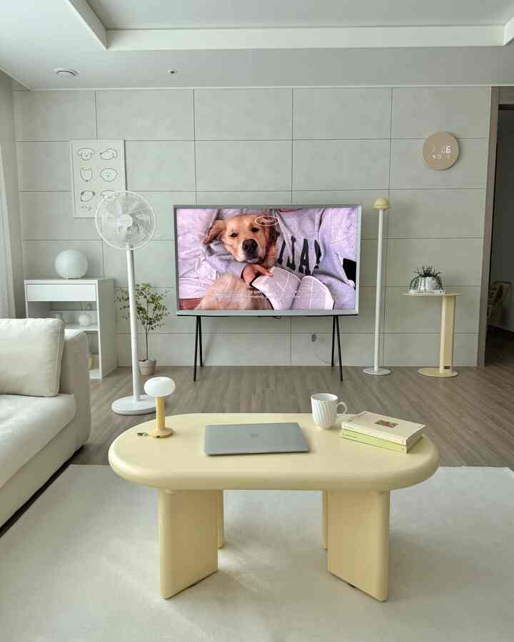 ホワイトとベージュのトーンのリビングルーム、シンプルなソファと黄色いテーブル、テレビスタンドが配置された清潔感のあるミニマルな空間