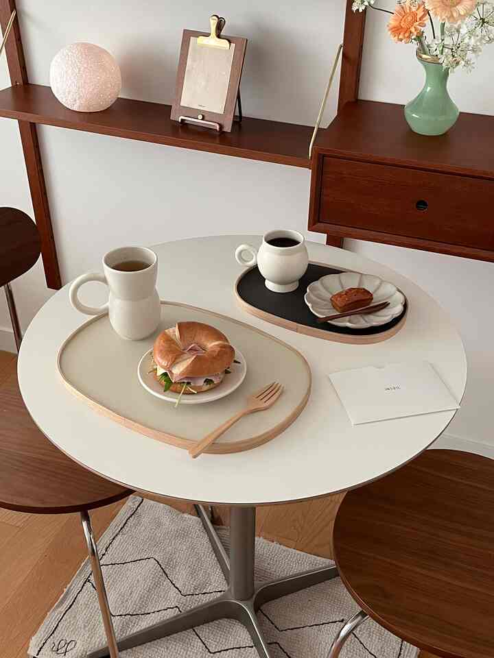 Bright white round table with brown wooden chairs, featuring natural tone mugs and trays holding bagel and dessert, creating a stylish home cafe space