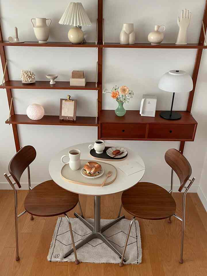 Natural brown and white toned dining room featuring a round dining table, wooden chairs, wall shelves, and ambient lighting