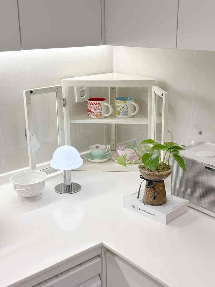 明るく清潔感のあるホワイト調のキッチンコーナーキャビネット、透明なアクリル扉とマグカップ、ティーカップ、小さな植物が配置された印象的な空間