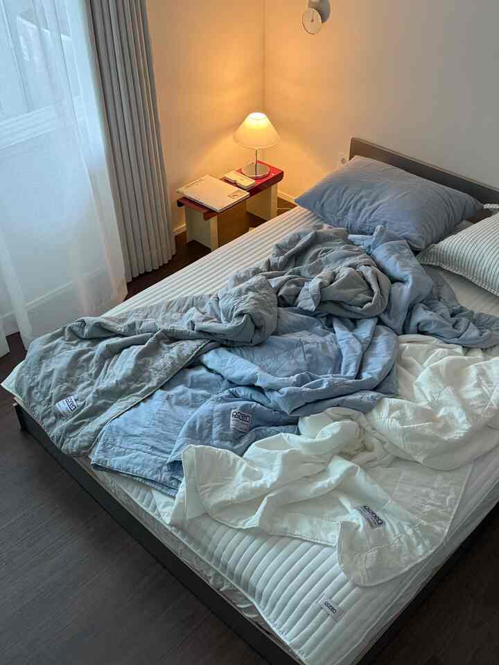 ナチュラルカラーのホワイトとブルーが調和するコンパクトな寝室、ベッドとサイドテーブル、カーテンが配置された雰囲気のあるインテリア