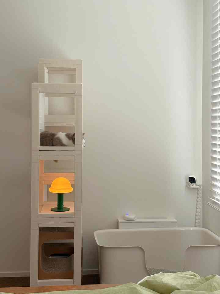 明るいホワイトトーンの寝室に、モジュール式ペット家具とくつろぐ猫、白い猫トイレ、木製ブラインドが配置された清潔感のあるインテリア