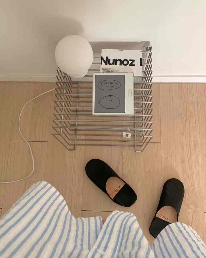 自然な木目調の床と白い壁の狭い空間、ワイヤー収納ラックの上にテーブルランプと電子書籍リーダーが置かれたミニマルなインテリア