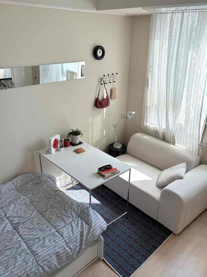 明るいホワイトトーンのワンルーム、ベッドとソファ、デスクが同じ方向に配置された落ち着いた印象的な空間