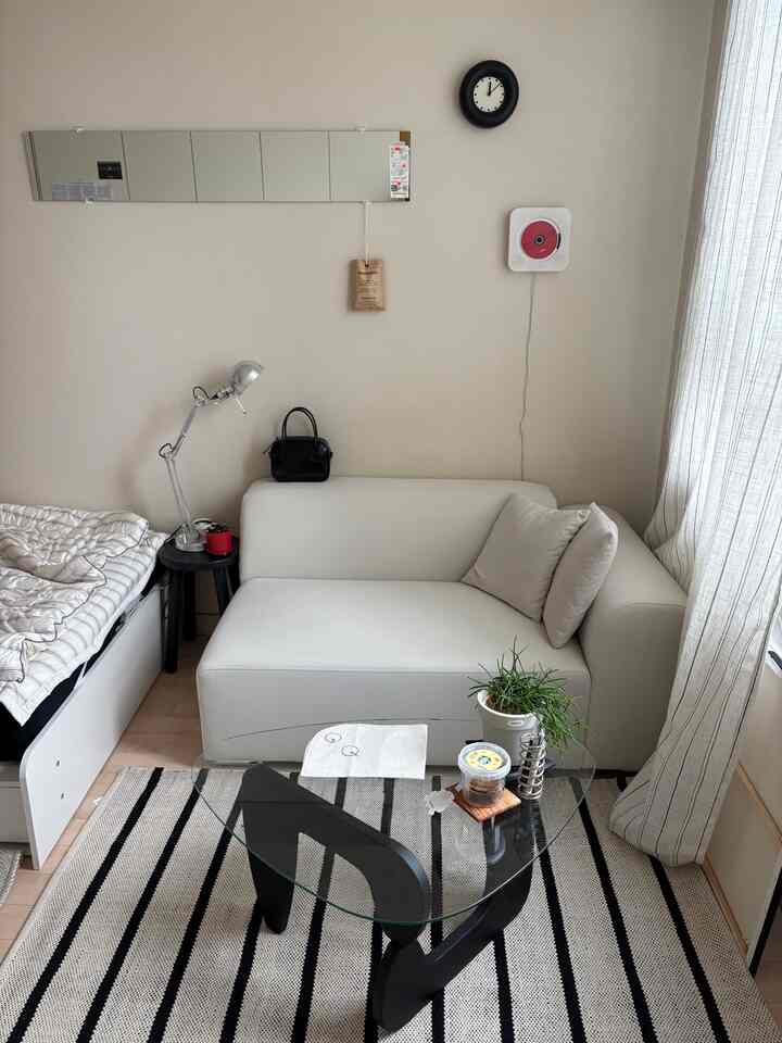 Bright natural-tone studio apartment living space featuring a white sofa, single bed, and striped curtains with a cozy atmosphere