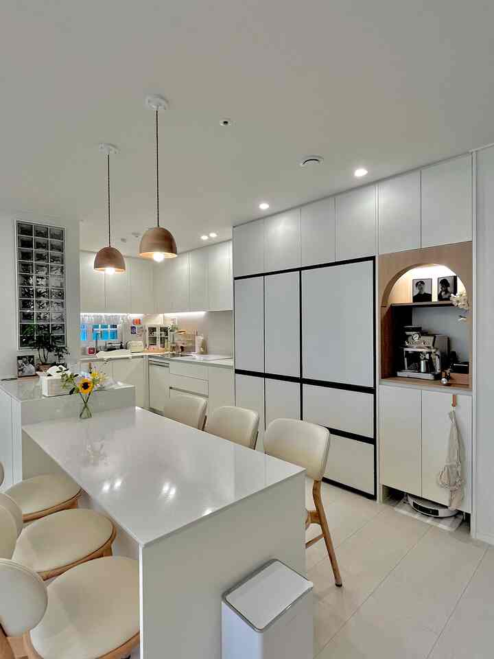 White-toned kitchen space featuring an island dining table, wood-tone pendant lights, and a cozy home cafe corner with a clean, natural interior