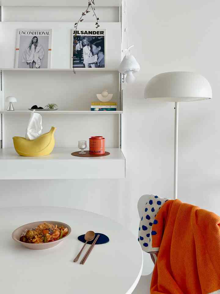 ホワイトトーンのリビング、丸いダイニングテーブルにオレンジのスローと照明が配置されたミニマルなインテリア