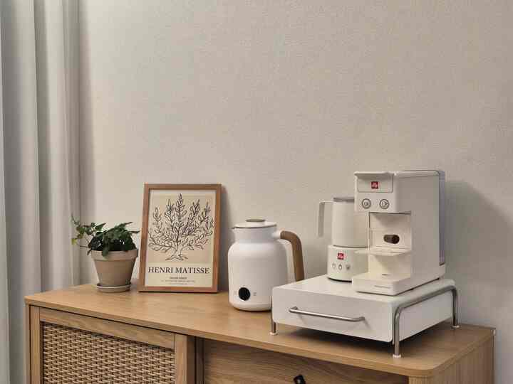 白と木目調のキッチン、電気ポットとコーヒーマシン、ポスターと植物が配置された雰囲気のある空間