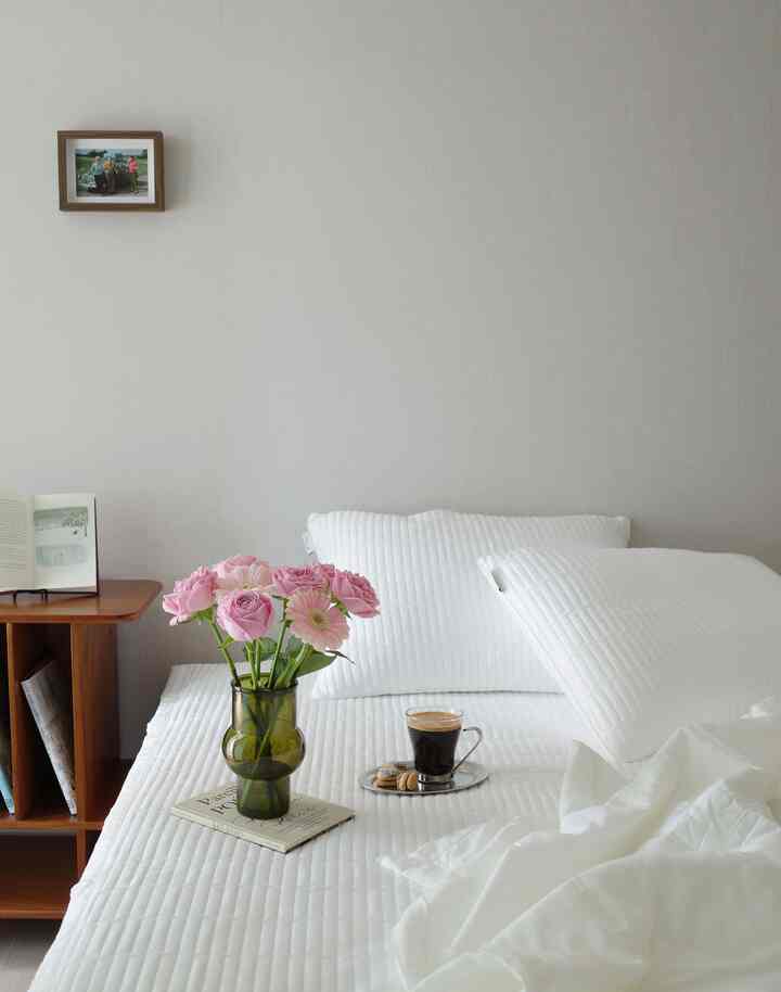 白を基調とした寝室、キルト寝具と枕、木製サイドテーブルにピンクの花瓶が置かれた落ち着いた空間