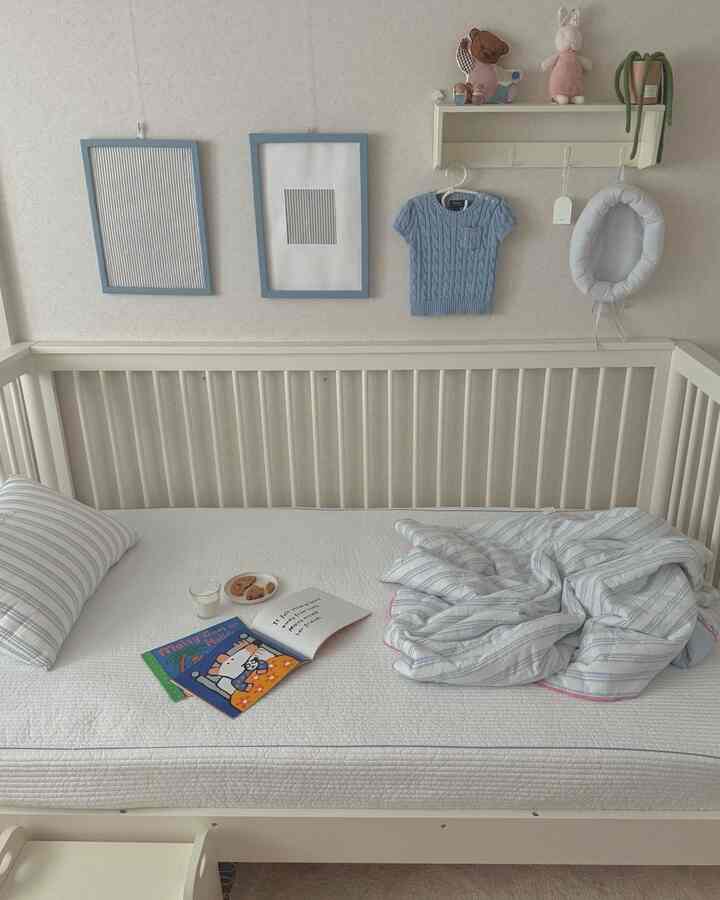 ホワイトとブルーの色調の赤ちゃん部屋、ストライプの寝具と額装されたファブリックアートが配置された落ち着いた雰囲気のインテリア