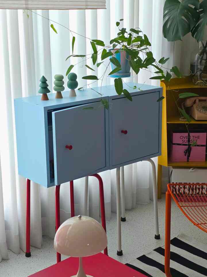 パステルブルーのサイドボードと白いカーテンがあるミニマルなリビング、赤い脚と緑の植物がアクセントの居心地の良い空間