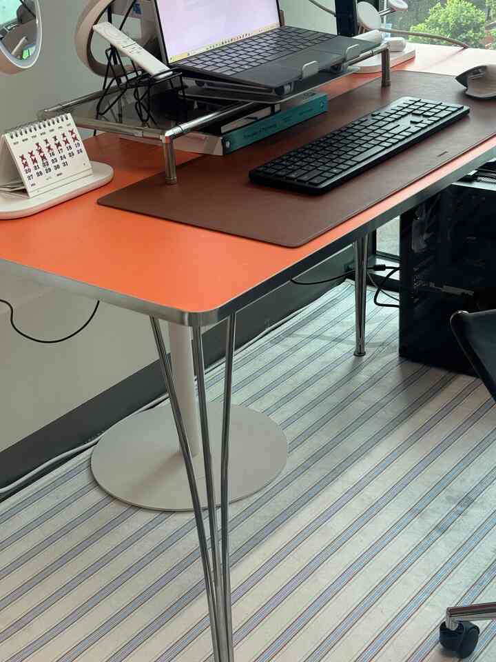 オレンジ色のデスクにモニタースタンドとレザーデスクパッドが配置された実用的なホームオフィス空間