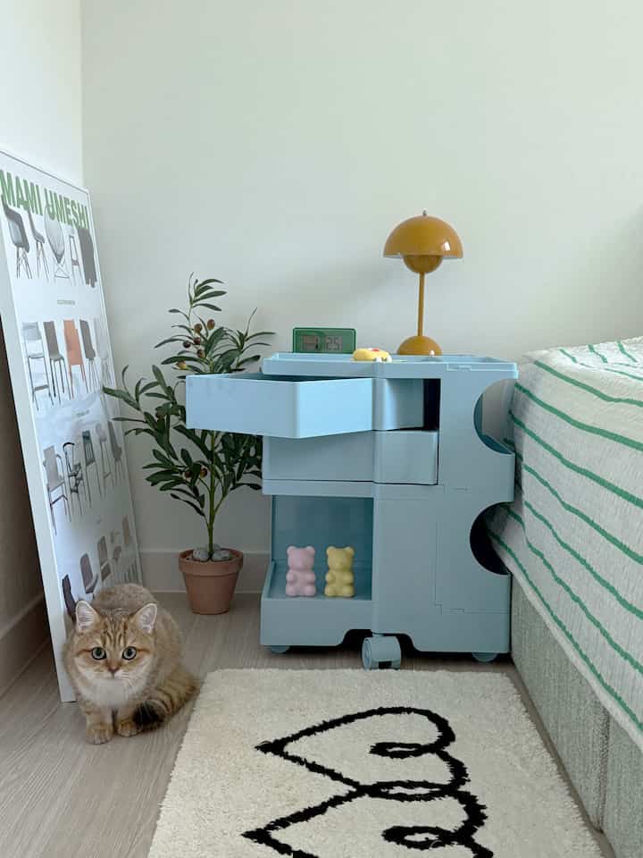Clean bedroom in white and mint tones featuring a rolling trolley beside the bed and a cat, creating a fresh vibe