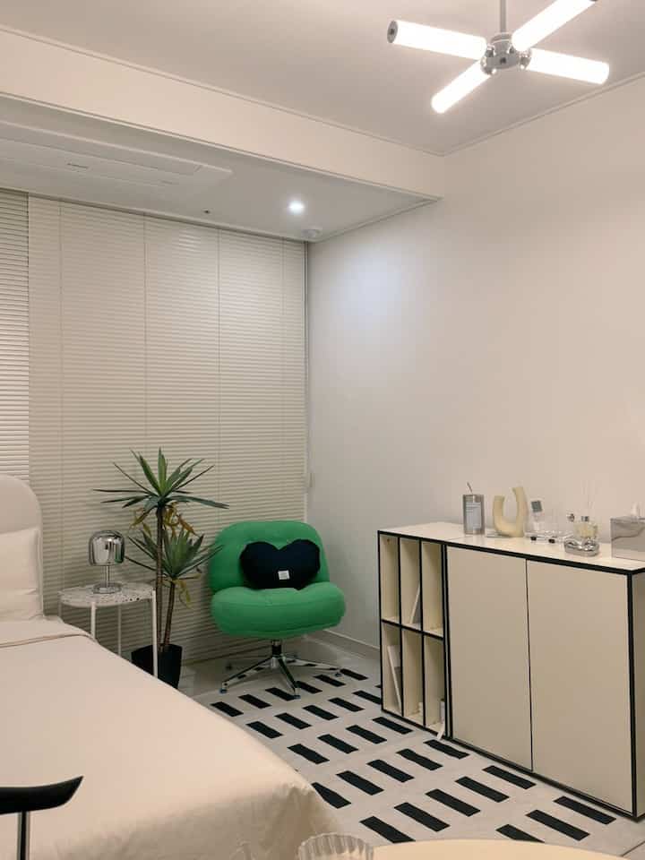 モノトーンとシルバー調のワンルーム寝室、ミッドセンチュリーモダンの緑色チェアとシーリングファン照明が配置された印象的な空間