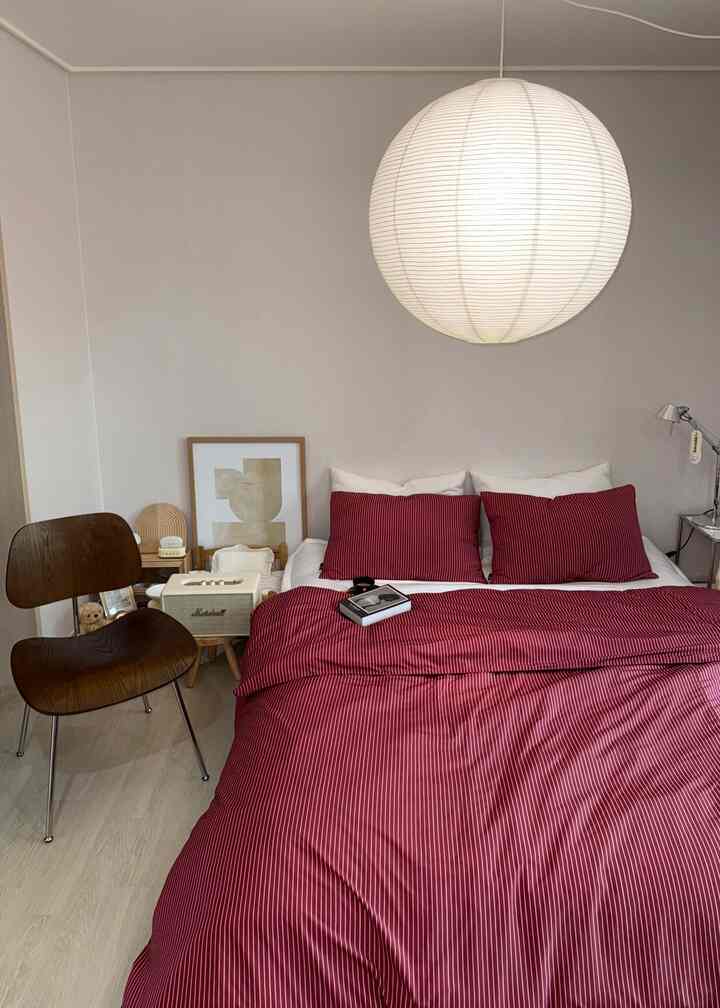 白い壁と天井が調和する寝室、赤いストライプの寝具が印象的で、木製チェアとテーブルランプが配置された落ち着いた空間