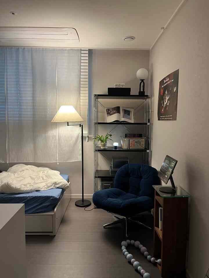 白い壁と木目調の床のワンルーム寝室、ネイビーのアームチェアとフロアランプが配置された落ち着いた一人暮らしの空間