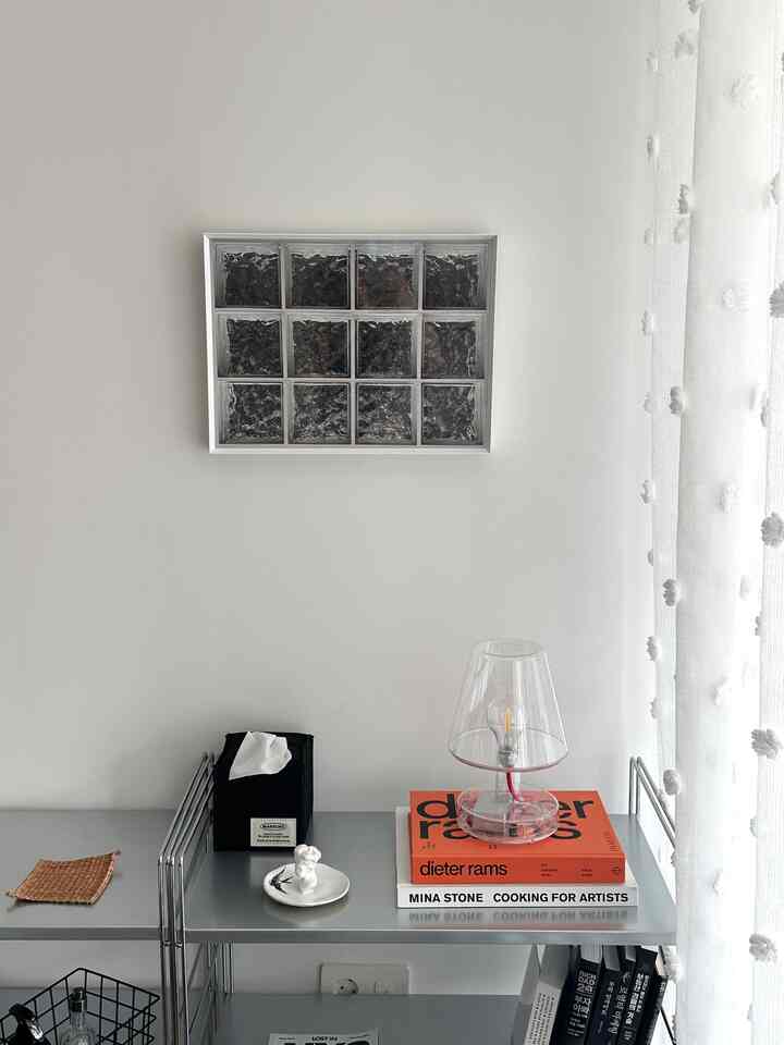 ホワイトとシルバーのトーンのモダンな空間、中央の壁にガラスブロックの額縁が掛けられ、その下にはスリムなメタルシェルフと透明なランプが配置された印象的なインテリア