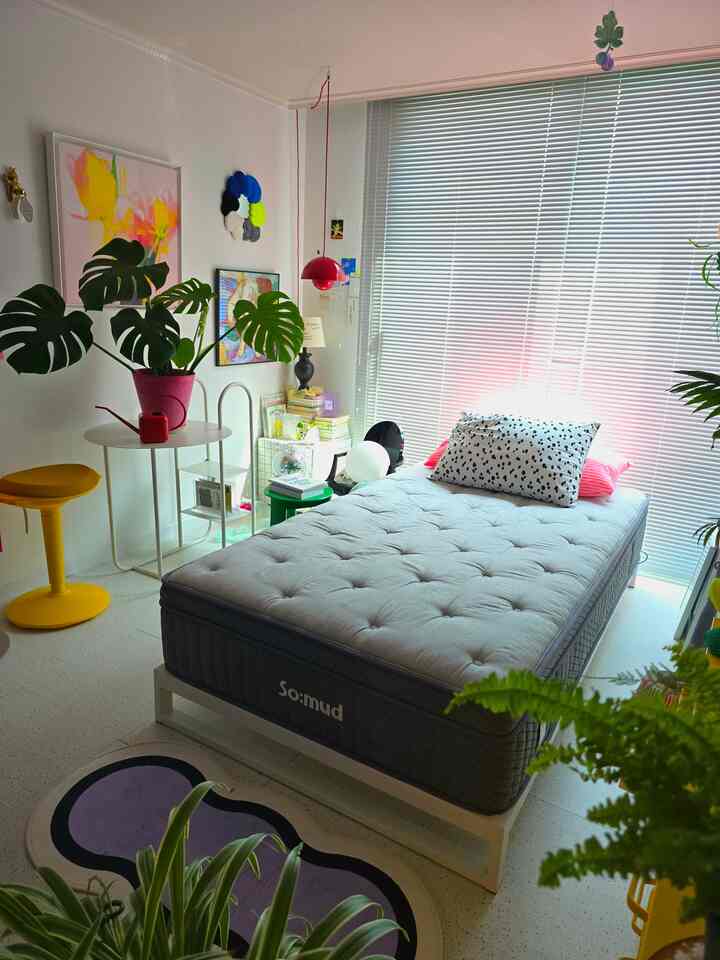 白と緑のトーンの寝室、中央にシンプルなベッド、多様な植物とカラフルなアートポスターが調和した雰囲気のあるインテリア