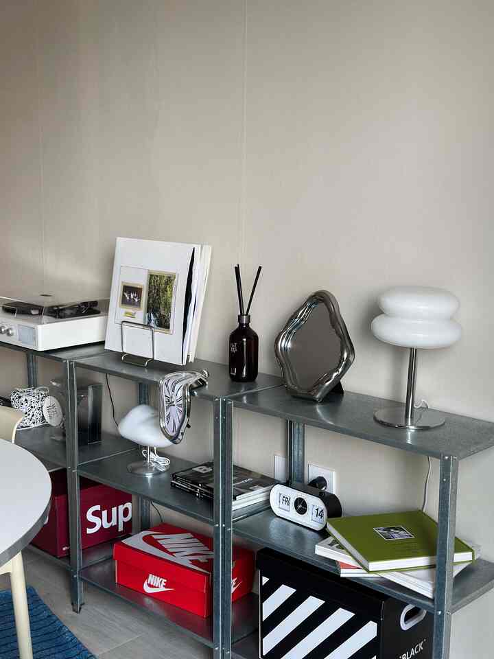 白い壁の前に金属製の棚があり、赤い箱や独特なデザインの卓上時計、ディフューザーが置かれたモダンな空間