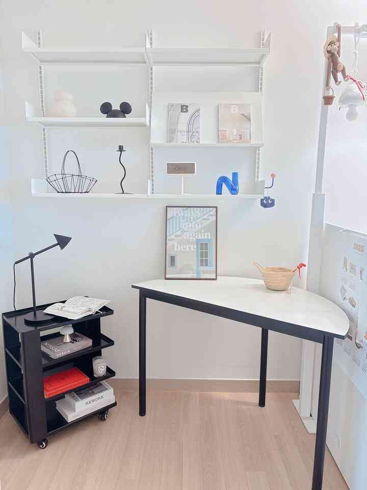 A simple, modern workspace featuring white walls and table contrasted by a black 4-tier mobile storage trolley