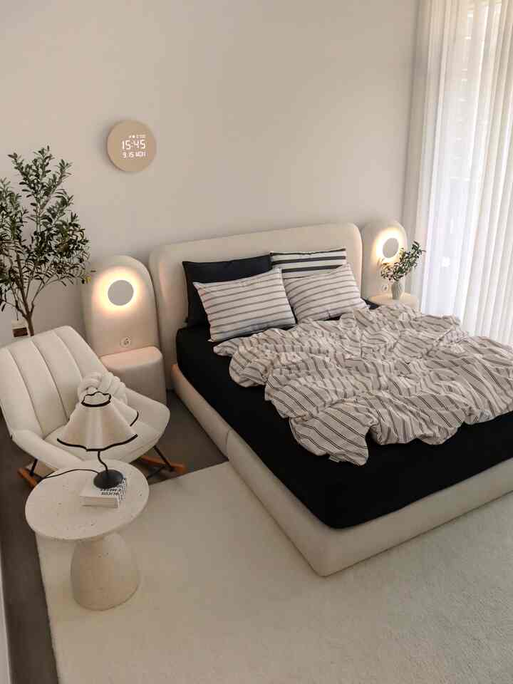 Black and white, neutral-toned bedroom featuring a fabric king bed, sheer curtains, olive tree, and rocking chair in a modern atmosphere