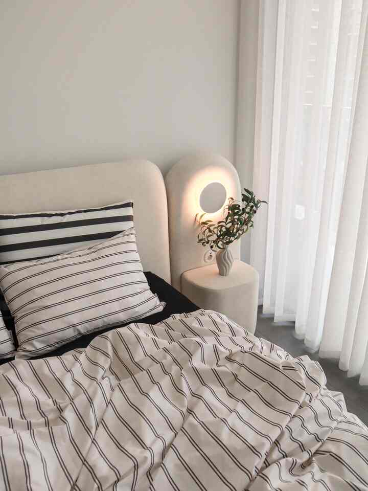 Bright natural-tone bedroom featuring a fabric bed with striped bedding, side table with table lamp and olive branch on the right, cozy atmosphere