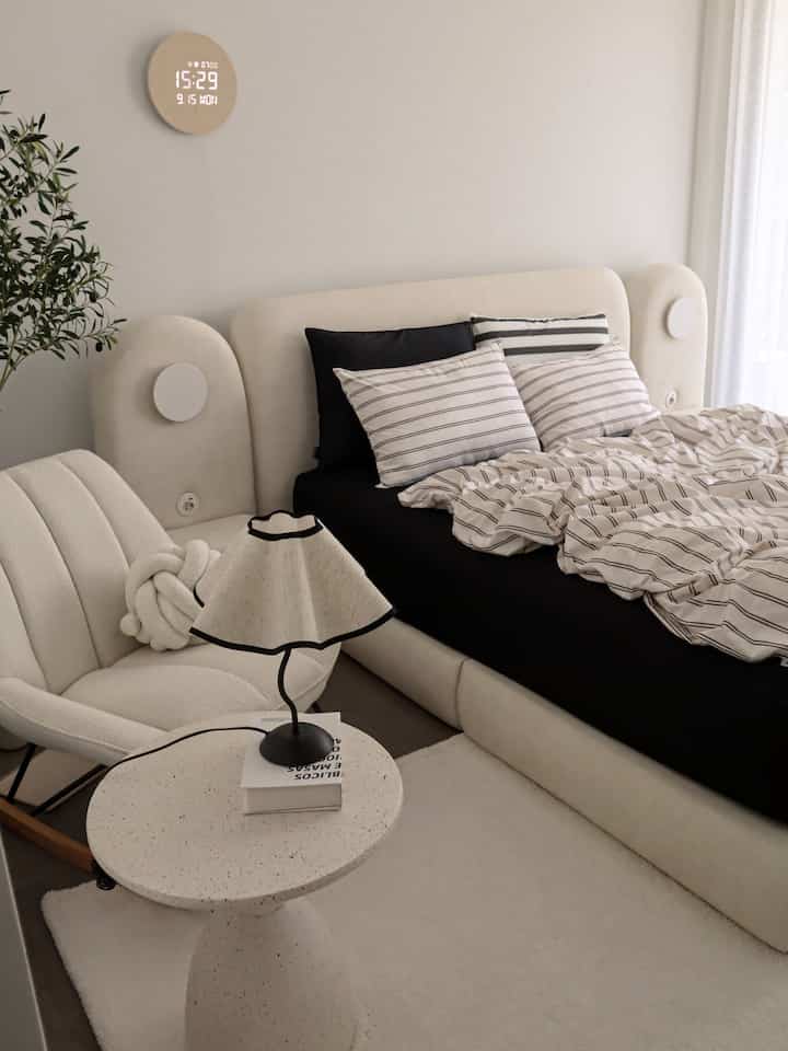 Neutral-toned bedroom featuring beige fabric bed, bouclé chair, striped bedding, and a table lamp in a modern setting