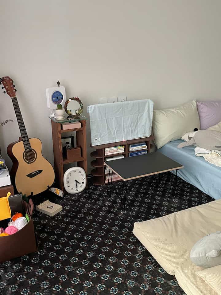 白い壁とパターンカーペットのある狭い部屋、ギターと本棚、床マットレスと小さなテーブルが配置された居心地の良い一人暮らしの空間