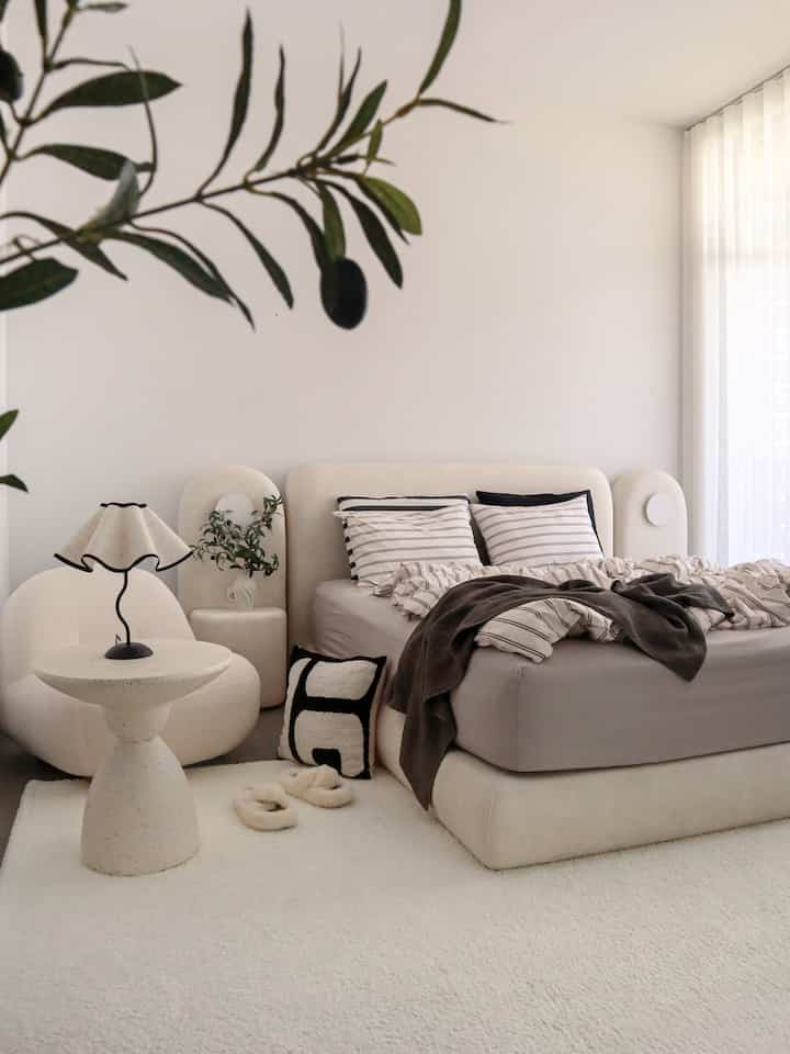 A bedroom in ivory and gray tones featuring a fabric bed, stool, and cushions in a simple natural modern setting