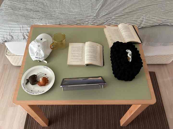 ナチュラルなブラウンの木製ダイニングテーブルにオリーブ色のランチョンマットが置かれ、ベッドと本とパンが自然に調和した雰囲気のある寝室空間