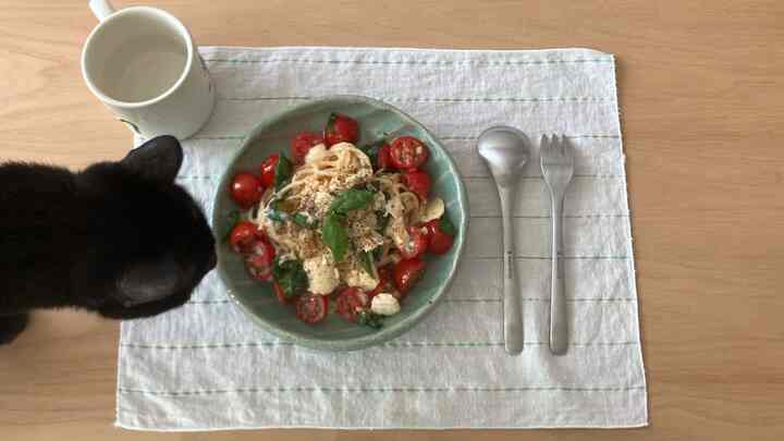 ナチュラルな木製テーブルの上にグリーンの皿と新鮮なパスタが置かれ、左側に黒い猫が覗く温かみのある食事空間