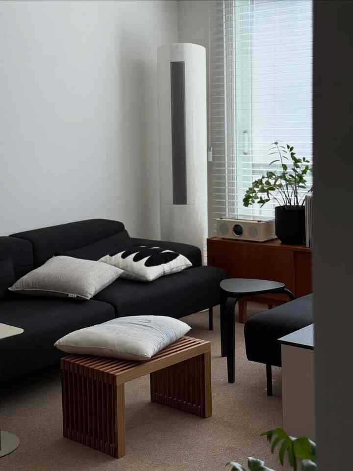 ホワイトトーンのリビング、ブラックのソファと木目調のベンチが配置され、明るいブラインドと植物が調和する落ち着いた空間
