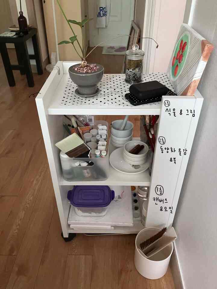 白と木目調が調和した狭い部屋のミニマルな収納キャスター付きカート、絵画用品と植物が配置された雰囲気のあるインテリア