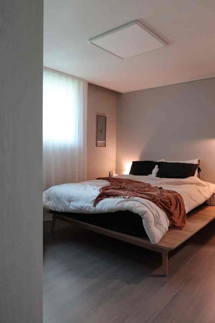 白とグレーのトーンの寝室、木目調のベッドフレームと柔らかなカーテンが配置されたモダンで落ち着いた空間