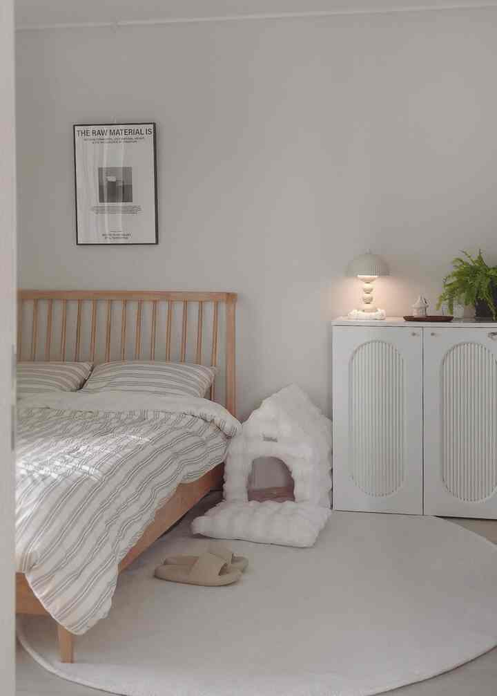 ホワイトとウッドトーンの寝室、木製ベッドと白いラグ、そして猫用の柔らかいペットハウスが配置された雰囲気のある空間