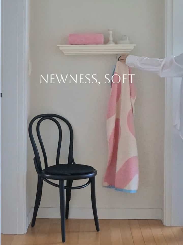 ピンクと白の色調の玄関、黒のダイニングチェアが壁際に置かれ、壁の棚にはピンクのタオル、柔らかな印象的な空間