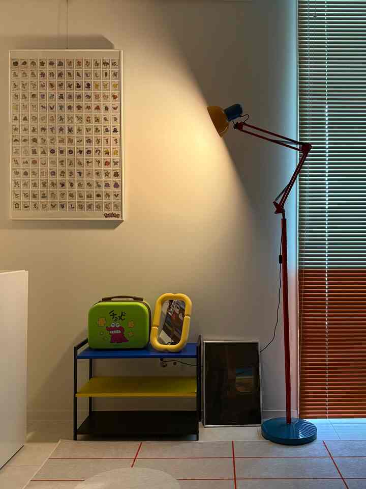 ベージュトーンの壁と床、ブラウンのブラインド、青と黄色の棚、赤いフロアランプが置かれたワンルームのインテリア