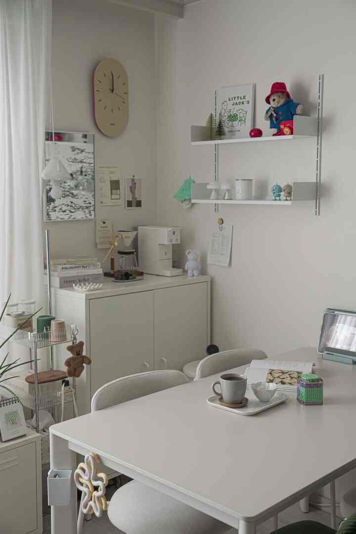 明るいホワイトトーンのダイニングルーム、中央に配置されたシンプルなホワイトのテーブルと椅子、壁の棚やキャビネットに小物が置かれた雰囲気のあるインテリア