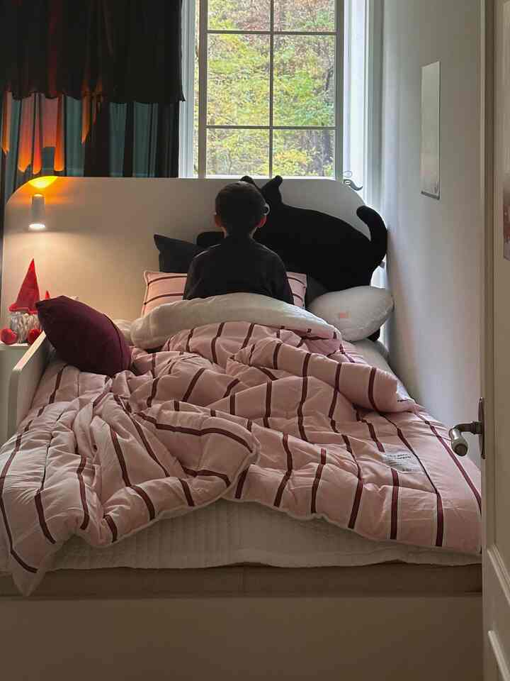 ピンクのストライプ寝具と黒いカーテンがある小さな寝室、子供がベッドに座り自然光が差し込む雰囲気のあるインテリア