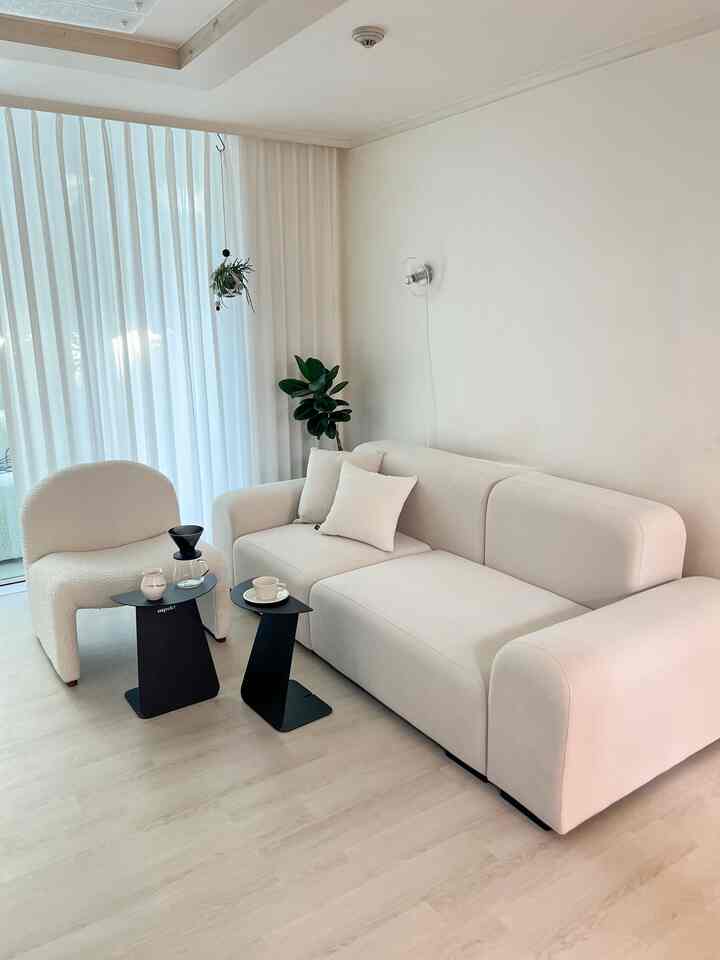 アイボリーとベージュのトーンのリビングに、左側に一人掛けのアームチェアと中央にブラックのサイドテーブル、右側に広いベージュのソファが配置されたシンプルで自然な雰囲気のインテリア