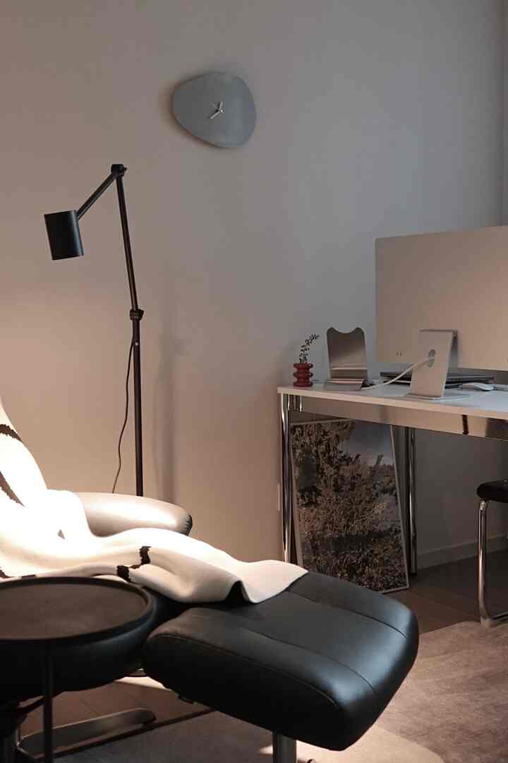 黒のレザーアームチェアと白いデスクが配置されたモダンでシンプルなホームオフィス、壁には独特なグレーの時計とフロアランプが置かれている