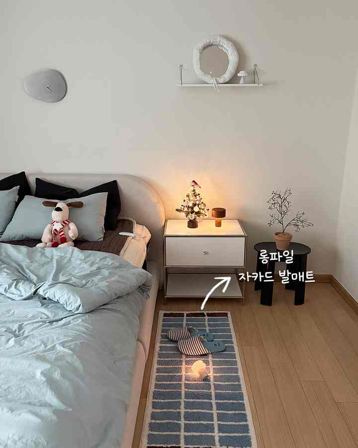 柔らかなブルーとナチュラルトーンの寝室、ベッド横にホワイトのナイトテーブルとブルーの玄関マットが配置された温かみのあるインテリア