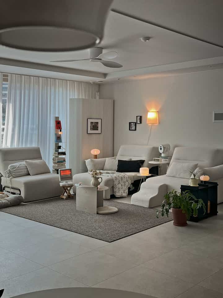 ナチュラルな色調のリビング、リクライナーソファとカーペット、ミニヒーターや多様な照明が配置されたシンプルな空間