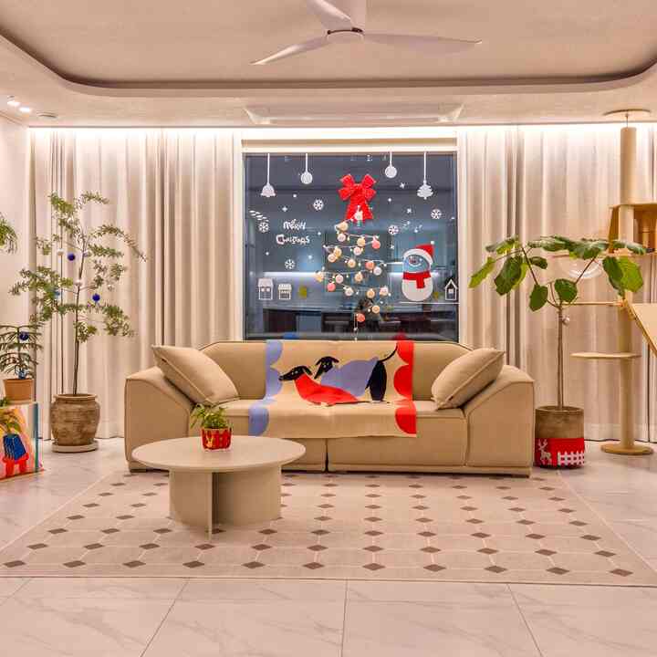 ベージュトーンのリビング、中央にベージュのソファと円形のコーヒーテーブル、両側に植物とキャットタワー、窓にクリスマス装飾が配された雰囲気のある空間