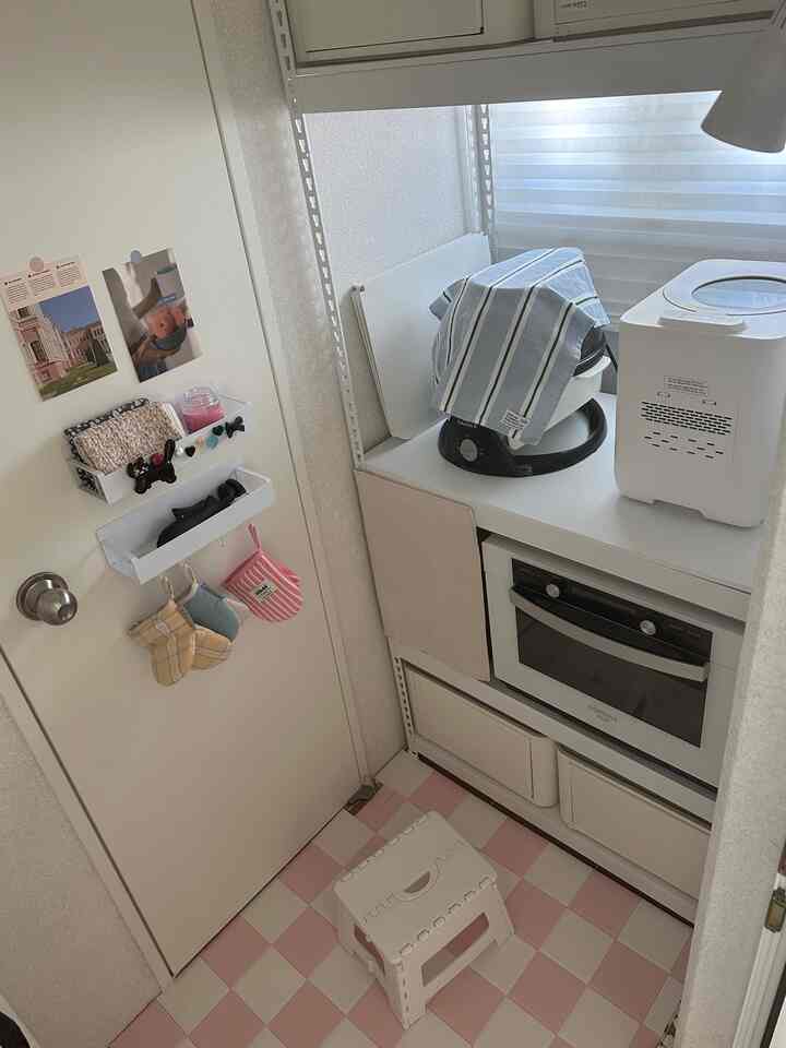 ホワイトとピンクの色調のキッチン、スピードラックの棚と小さなスツール、ブラインドが整然と配置されたコンパクトな空間