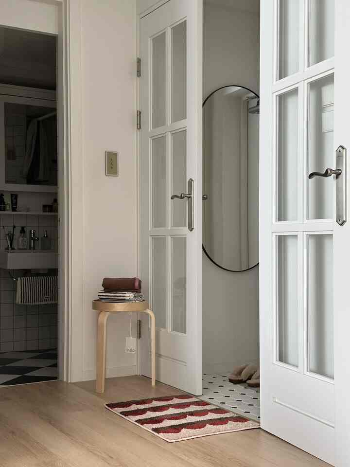白い壁とフレンチドア、木目調の床が調和した玄関、スツールにタオルが置かれ、大きな鏡が壁に掛けられた清潔で落ち着いた雰囲気のインテリア
