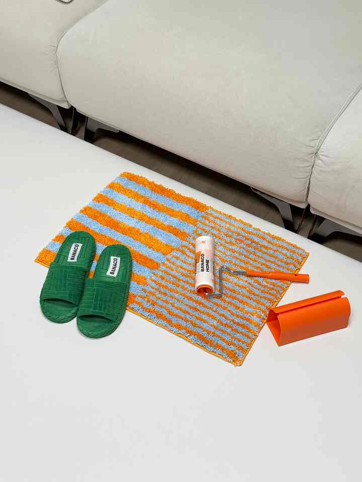 白いソファと緑の室内履きがあるリビング、オレンジと青のストライプのバスマットと掃除用具が配置された雰囲気のあるインテリア