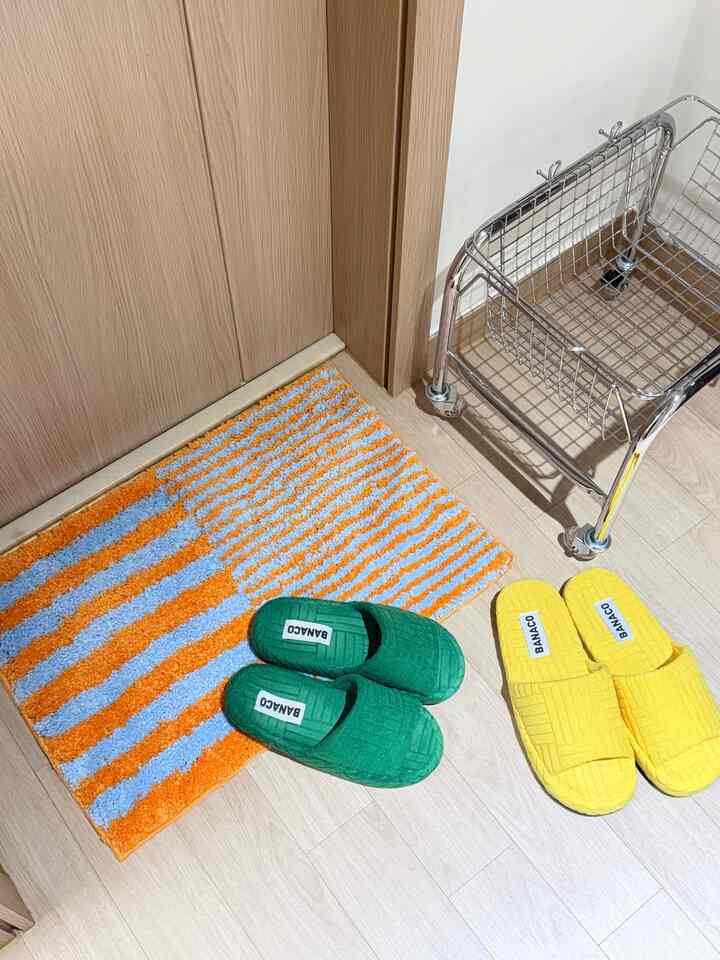 ナチュラルな木目調フローリングの小さな玄関スペースに、鮮やかな黄色と緑のルームシューズ、オレンジと青のバスマット、ステンレスのキッチンワゴンが配置された印象的な空間