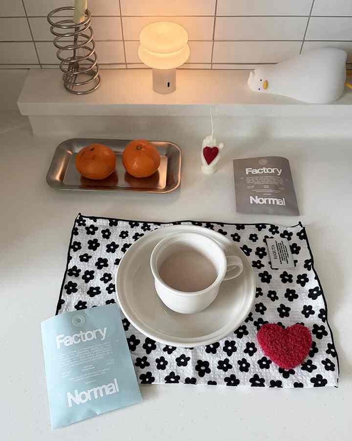 ホワイトとブラックの花柄テーブルランナーが敷かれたキッチンテーブルに、皿とマグカップ、香りのサシェが配置されたミニマルで清潔感のある空間