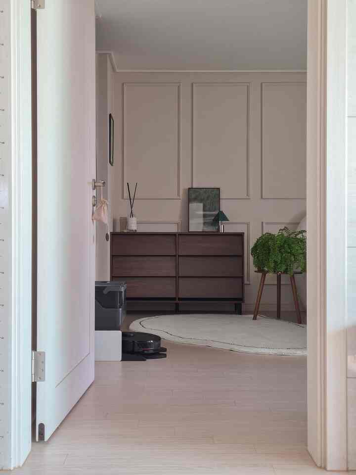 落ち着いたナチュラルトーンの寝室、木製のドレッサーと白いラグ、緑の植物が配置された雰囲気のあるインテリア